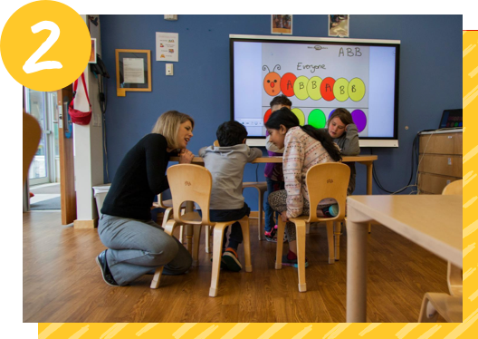 A group of children sit around a small table in a classroom at the Ronald McDonald House in Toronto. Their teacher is kneeling down next to the table.